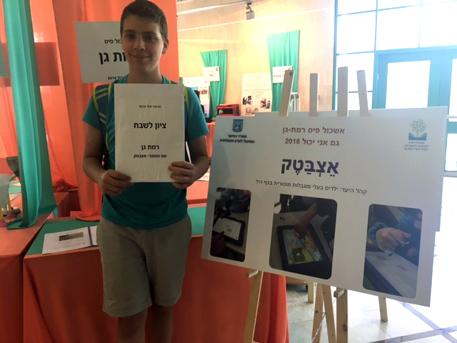 תלמיד אוחז בתעודת ציון לשבח ליד פוסטר המתאר את פרויקט אצבטק